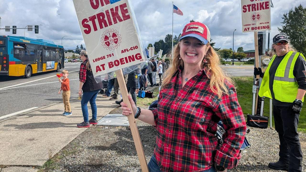 A smiling Kerri Foster wearing a red checked shirt and baseball cap, holding a strike sign outside of Boeing, with the American flag seen behind her and a bus going past on the road