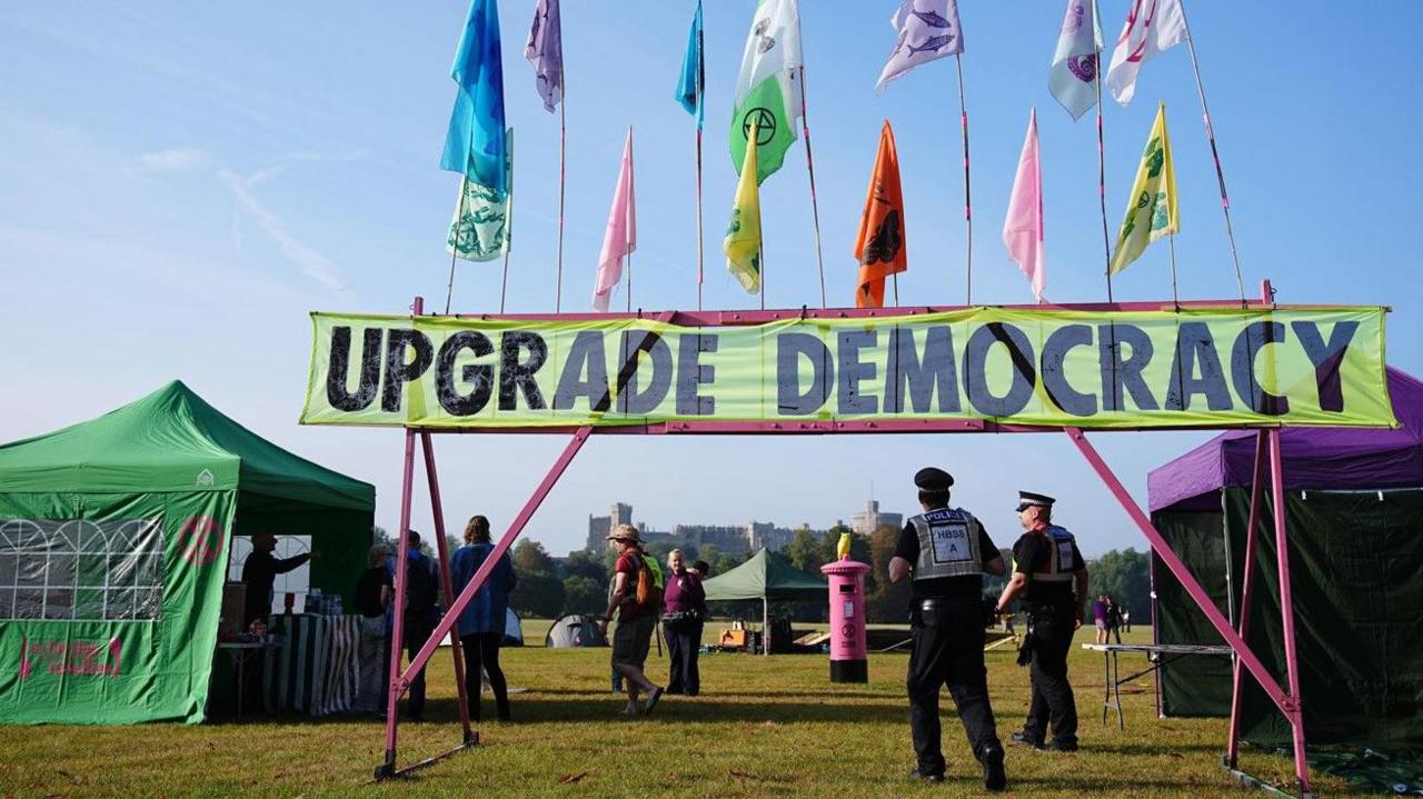 Two police officers walking beneath a metal frame holding a banner which says Upgrade Democracy. There are a number of tents and gazebos pitched nearby. Windsor Castle can be seen in the distance