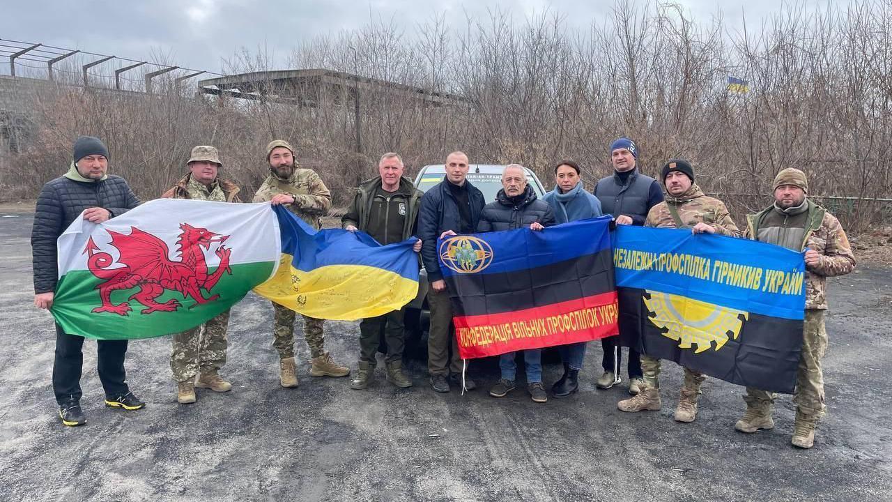 A group of men stood in a row behind four different flags, including a wales flag and a Ukraine flag. The other flags are black, blue and red, and black and blue, and contain Cyrillic writing. 