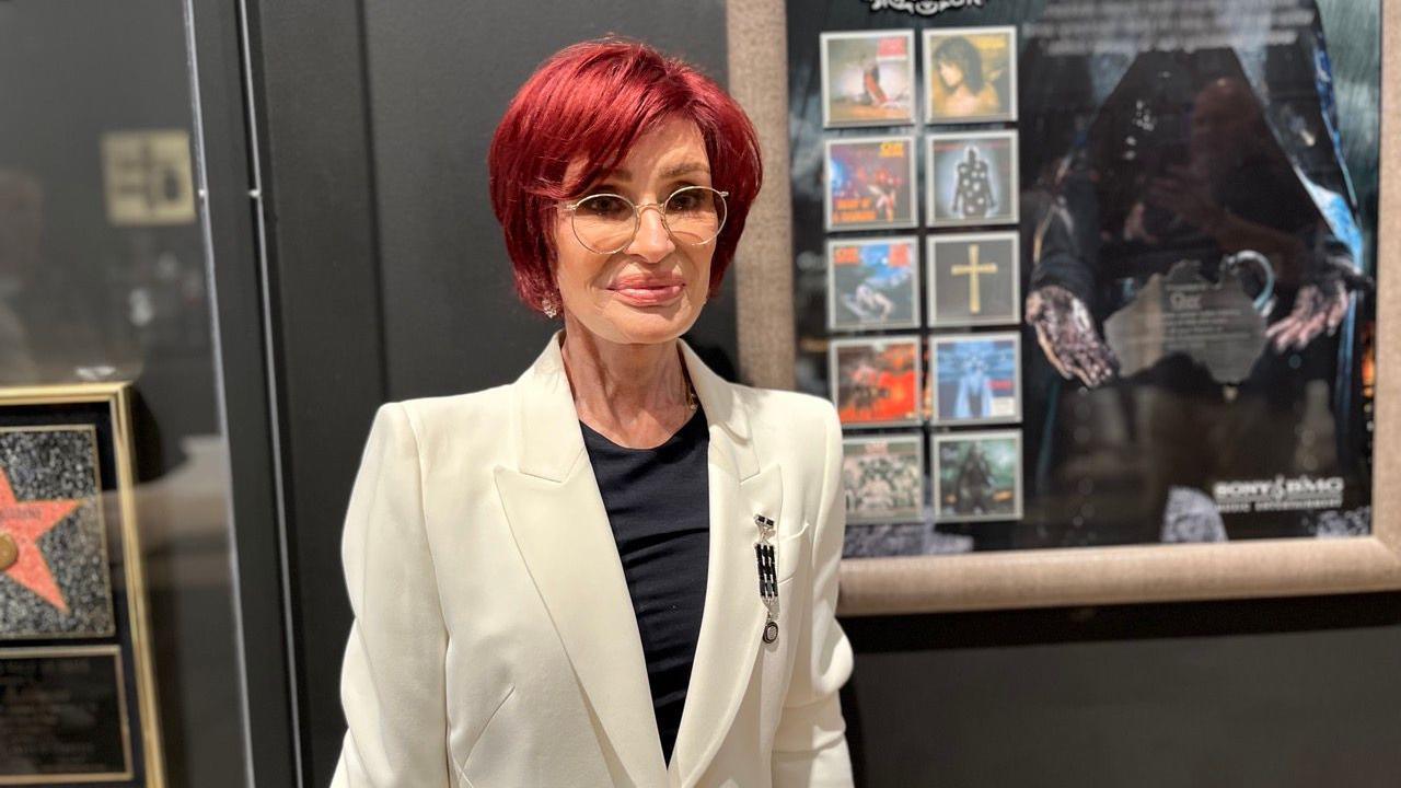 A woman stands in front of framed pictures on a black wall. She has short dyed red hair and glasses. She is wearing a white suit jacket and a black top.