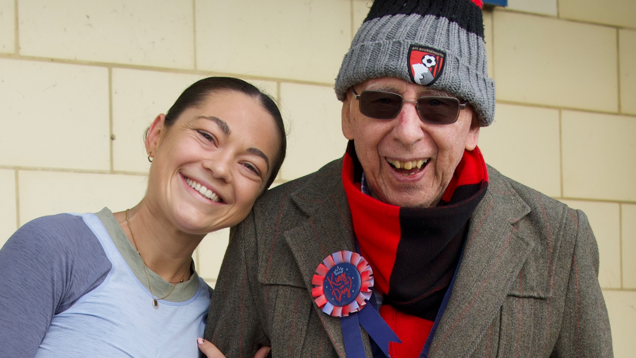 Issy Sedgwick with her grandfather, Ivor, smiling for the camera at the end of her challenge. She is wearing a grey running top and her hair is tied back. He is wearing a black and red AFC Bournemouth woolly hat and scarf and a brown coat with a blue and red rosette on the lapel.