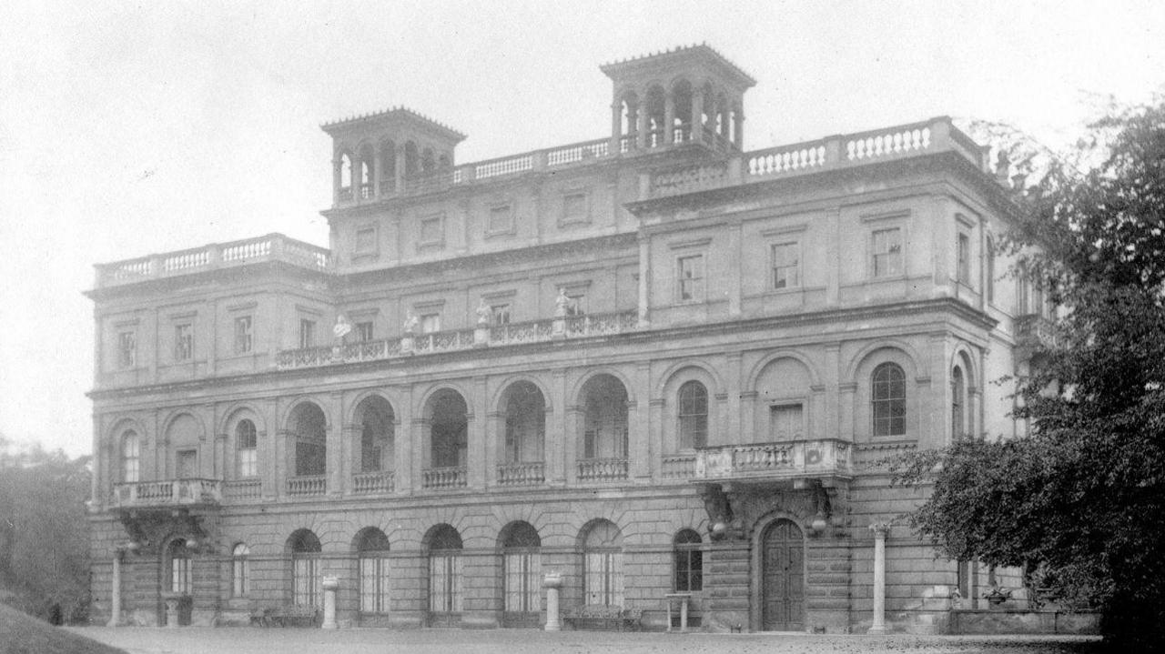A black and white external image of Deepdene House, a four-storey neo-palladian house in the country, with arched windows, two first-floor balconies and two turrets.