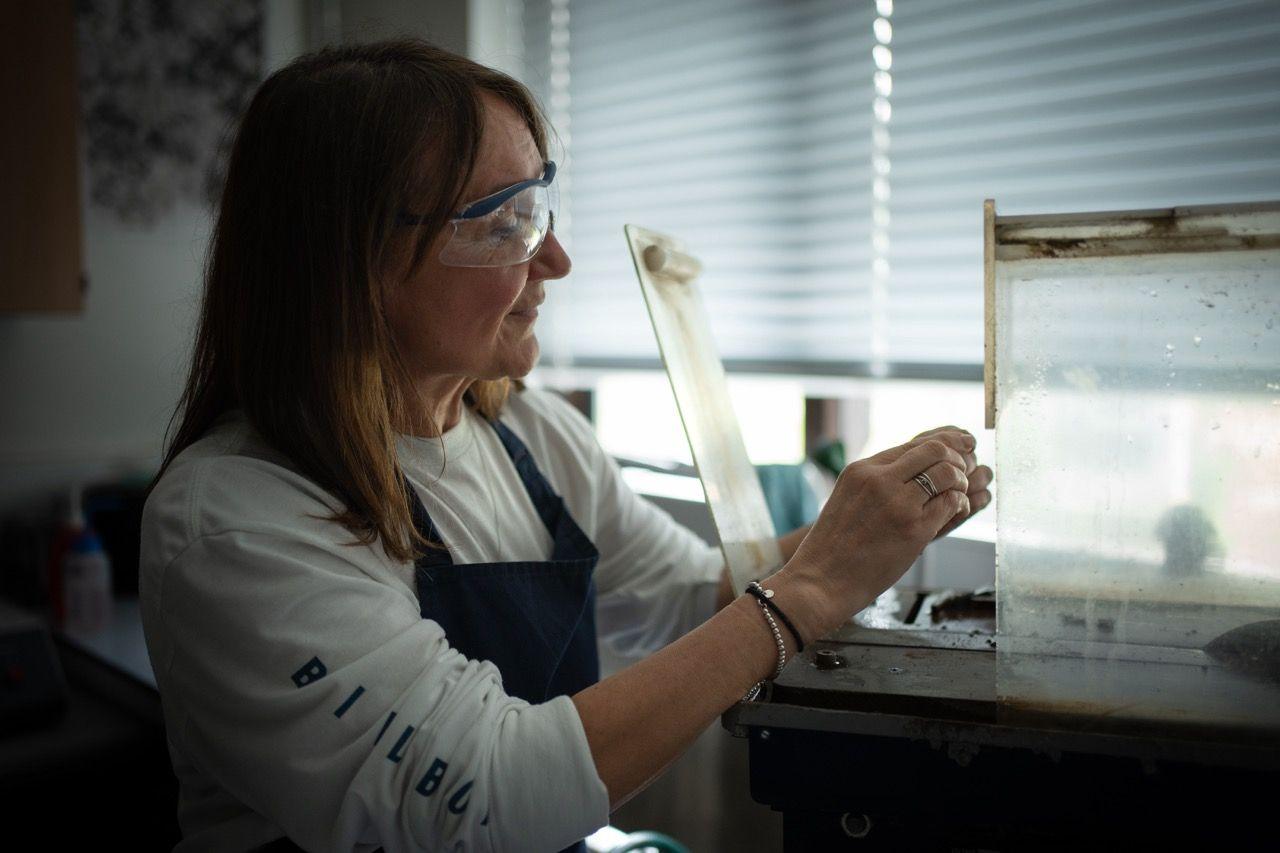 Kay Knight stands with her arms oustretched and hands above a metal surface. She is wearing white safety glasses. In front of her is a machine with perspex glass.