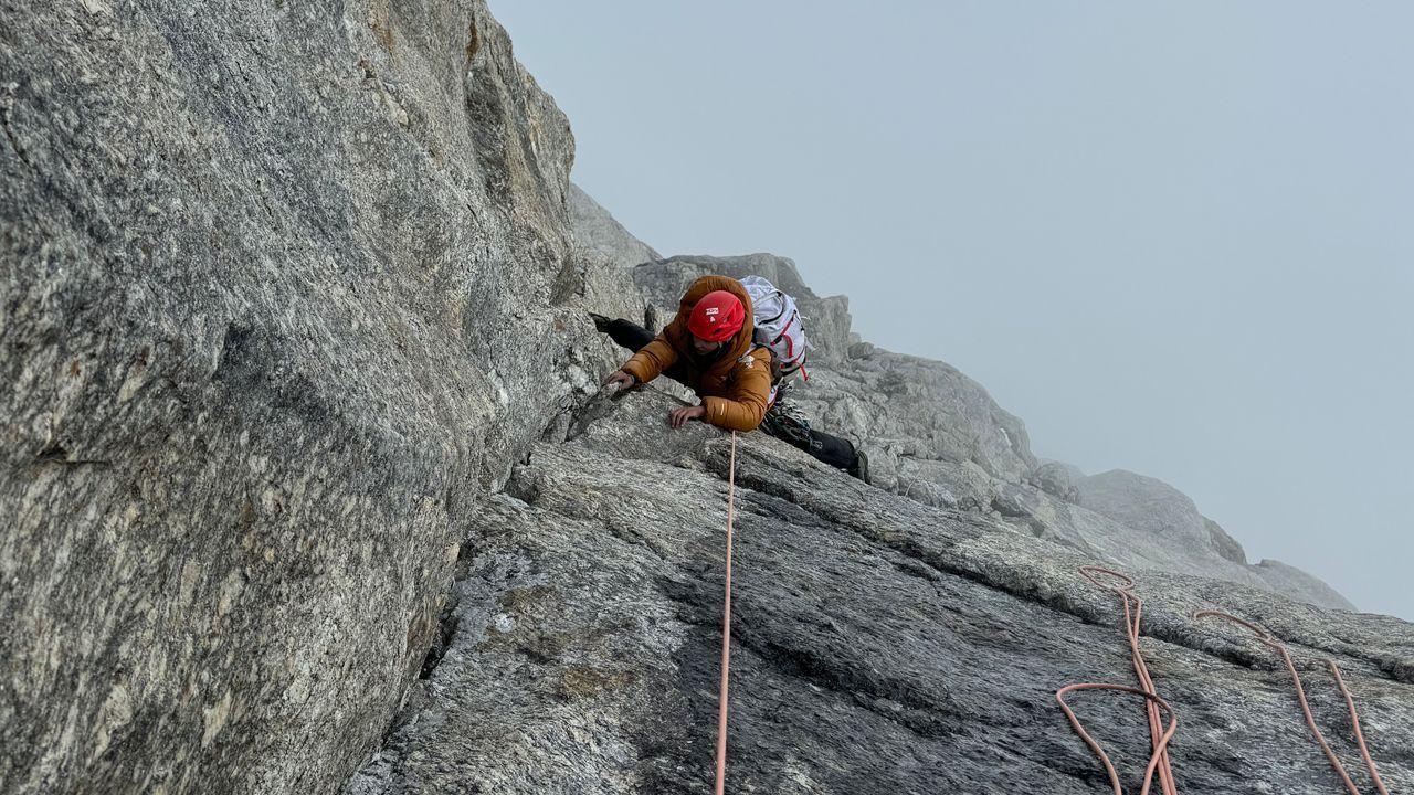 Taken from above, a woman in orange, her face obscured, is climbing the sheer face of a mountain, below her is thick fog obscuring the drop.