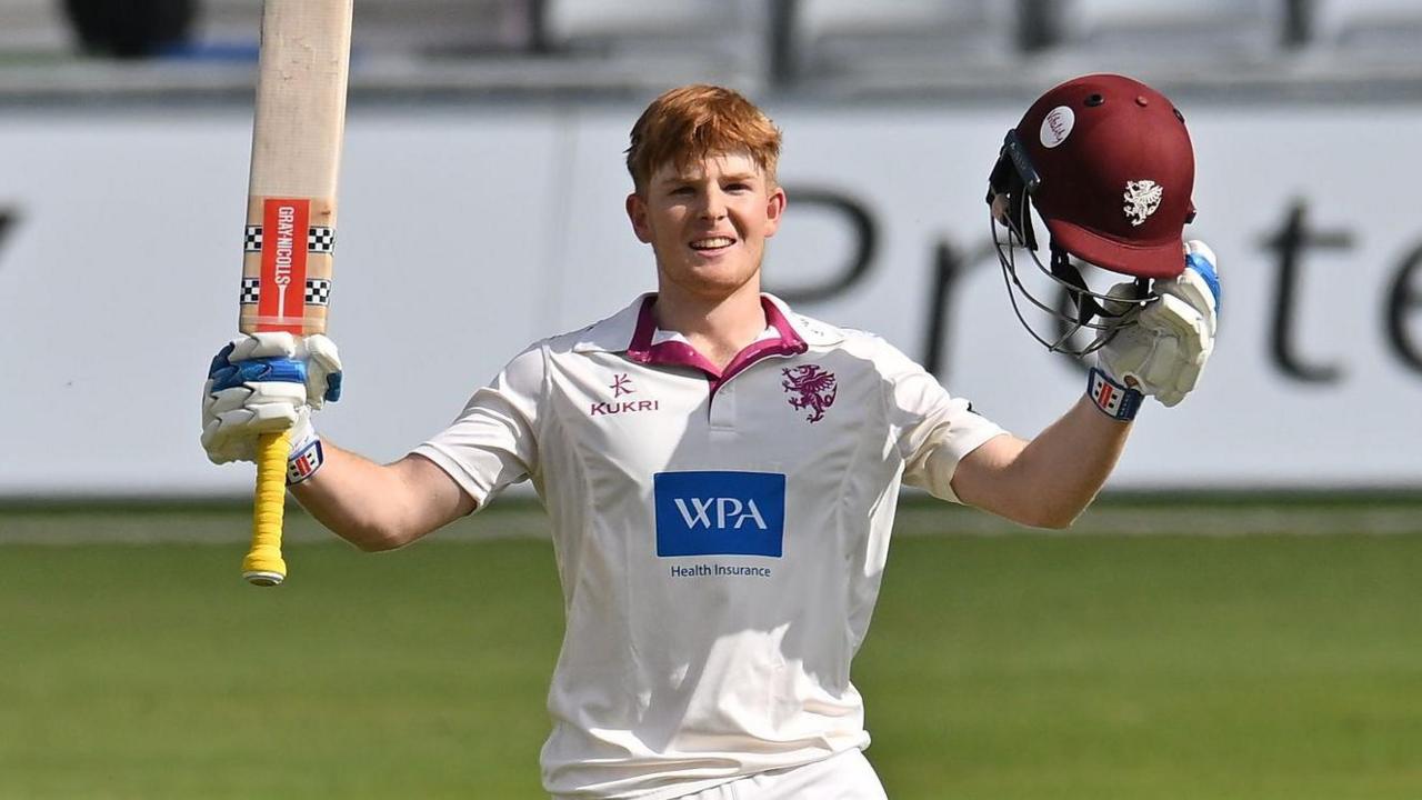 Thomas Rew raises his bat and helmet to celebrate scoring his maiden century
