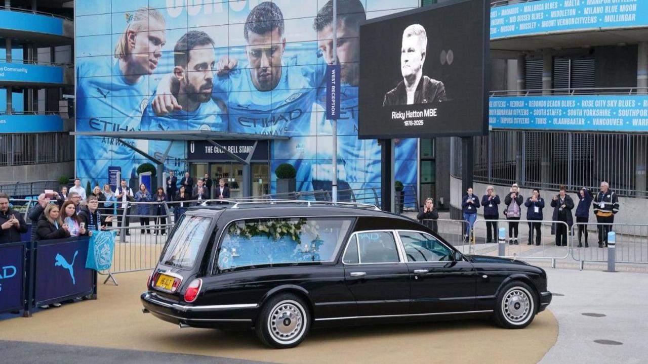 The black hearse is parked up outside The Etihad Stadium. A black and white photograph of Hatton is on the screen in front of the stadium.