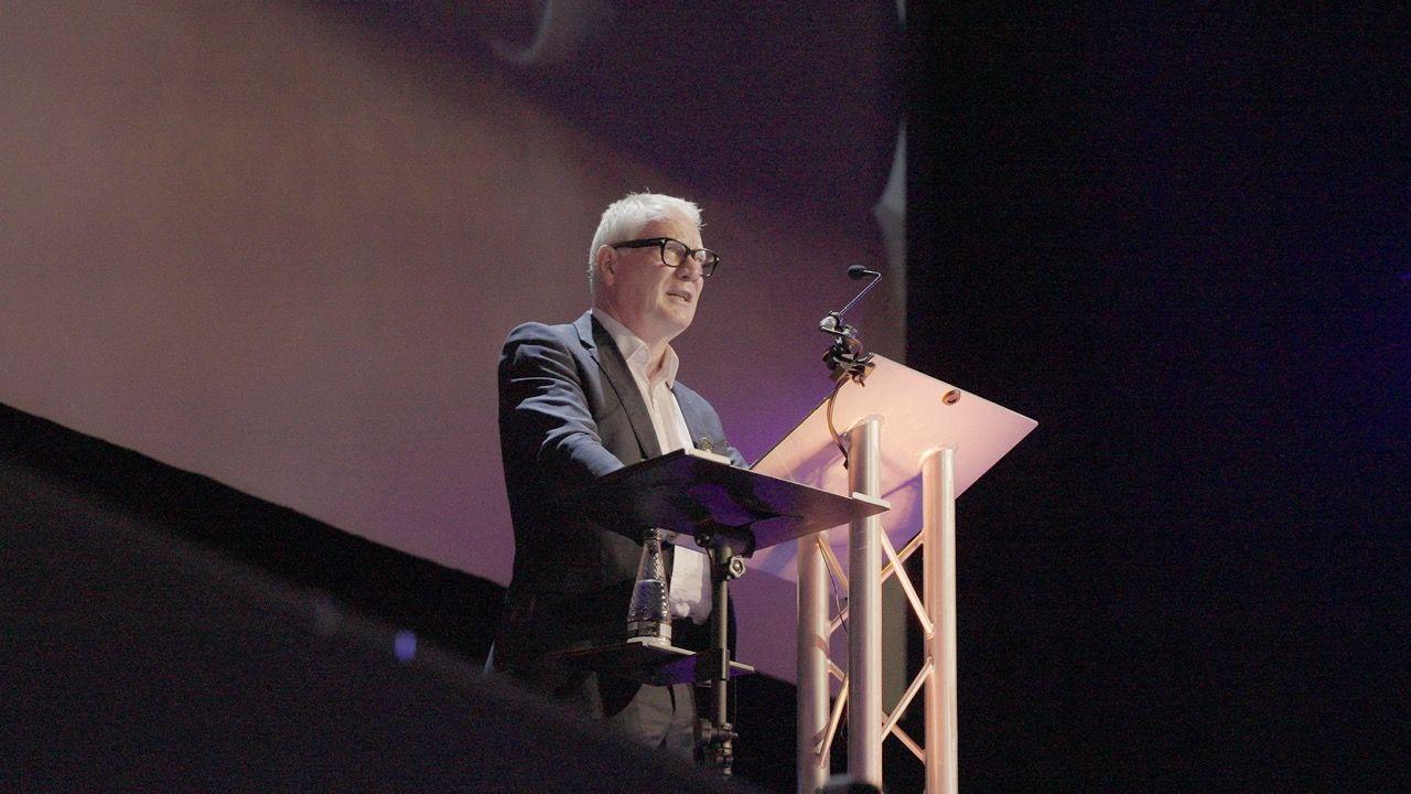 Mayor Richard Parker wearing a grey suit and white shirt, standing on a stage on a podium speaking into a microphone