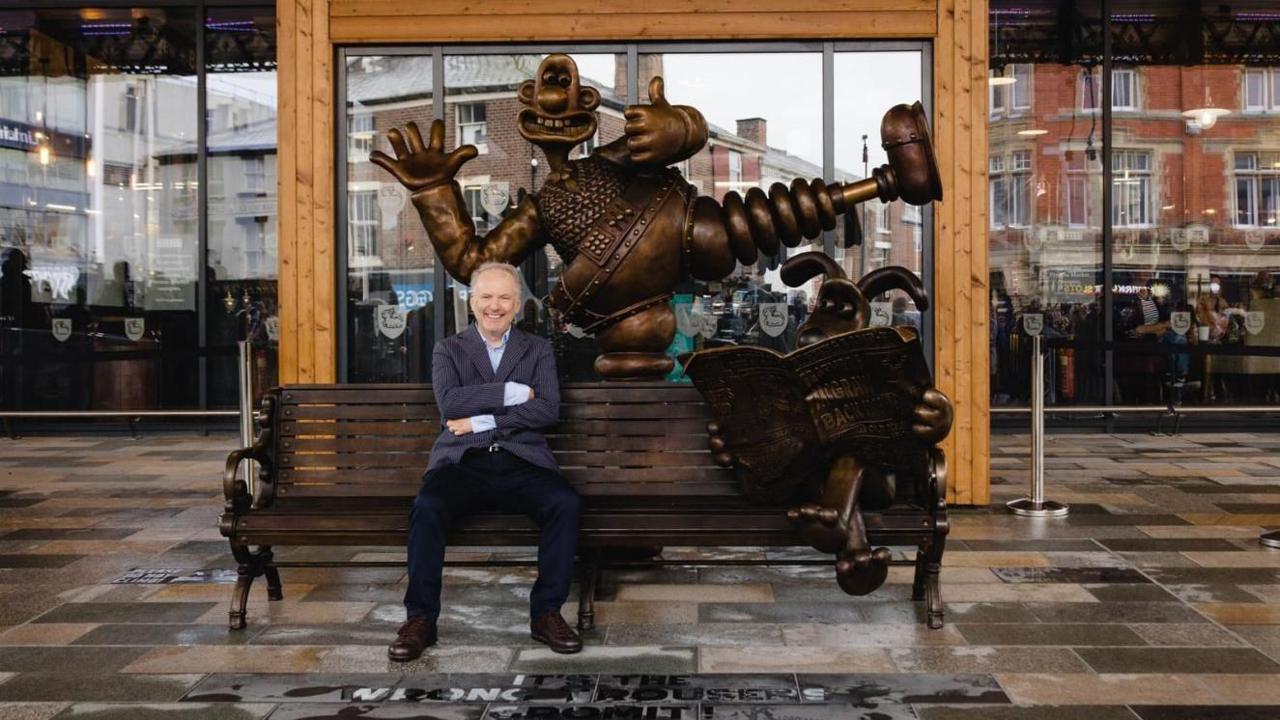 Nick Park sits on the bench that forms part of a statue of Wallace and Gromit. He wears a blue jacket, and smiles at the camera with his arms folded. The bronze-coloured statue includes Wallace, who stands behind the bench in "the wrong trousers". Gromit sits on the right side of the bench reading a newspaper.  