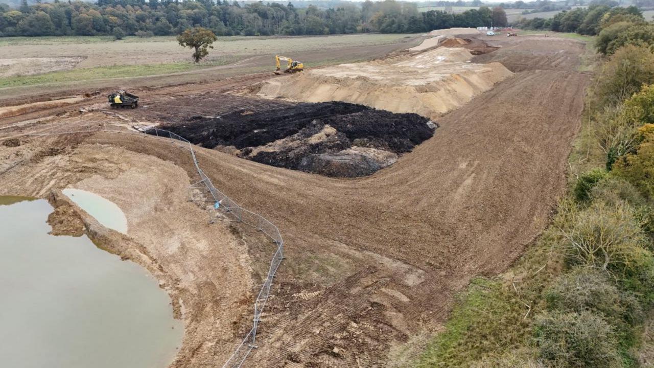 Groundworks to the side of the road, with trees on the right-hand side of the image and a large pool of water fenced off.