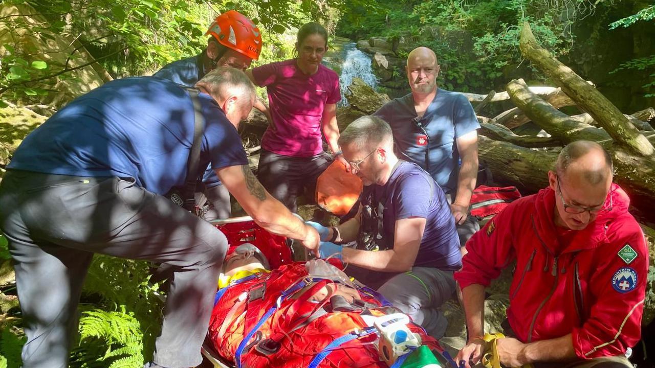Five people surround another person strapped to a stretcher. One is wearing a helmet, another is wearing a jacket with a mountain rescue logo on it. They are outdoors in an area full of greenery and fallen trees.