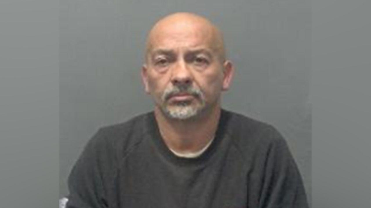 A police mugshot of John Angeli, who is bald with a grey goatee. He is wearing a grey top.