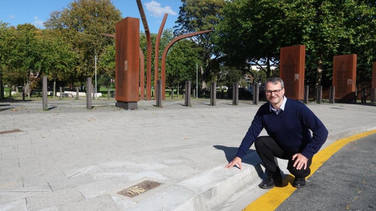 A man is pictured crouching near a curb marked with a thick yellow line, he rests his left hand on this. He is in front of tall rust-colored metal sculptures in a park-like setting under a blue sky. An outdoor scene featuring a person crouching near a curb marked with a yellow line. The man is wearing black glasses, a blue jumper, and black trousers.