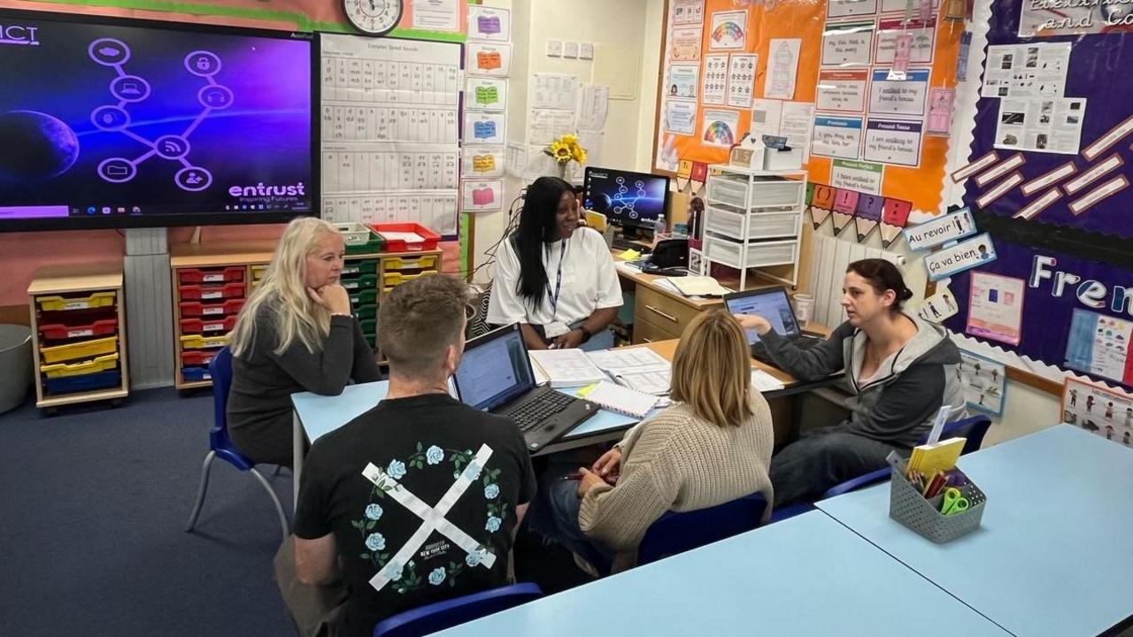 A group of five teachers sitting around a blue table in a classroom. A laptop sits on the table and the walls are filled with educational content