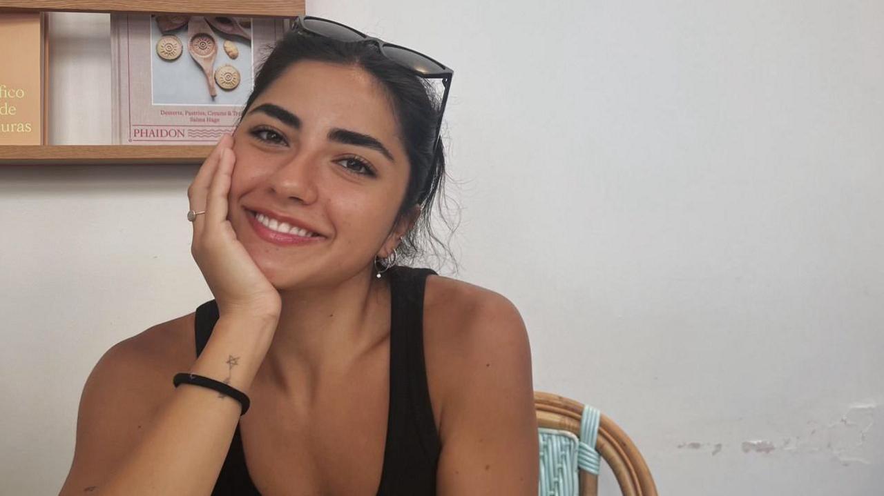 A girl with dark hair, tied up smiling at the camera. She is wearing sunglasses on her head and a black top, sitting in a cafe. In the background, there is a shelf with books. 