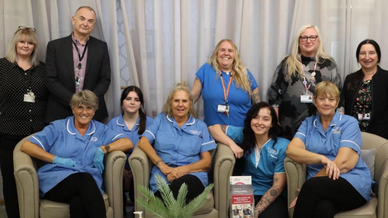 Staff from Davies Court Care Home, five, smiling female uniformed members of staff are seated or squatting in the foreground.  In the background are another four members of staff, standing. the group includes one male staff member, and Rotherham Councillor Joanna Baker-Rogers.