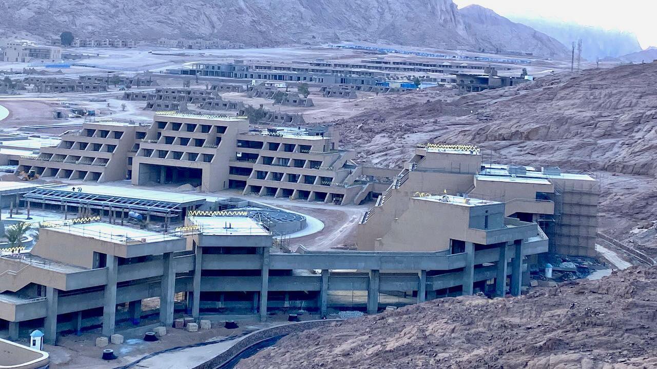 The hotel building still appears to be mostly a shell, and around four storeys high. Smaller buildings, also in the valley and still under construction, can be seen in the background, with the surrounding mountains in the background