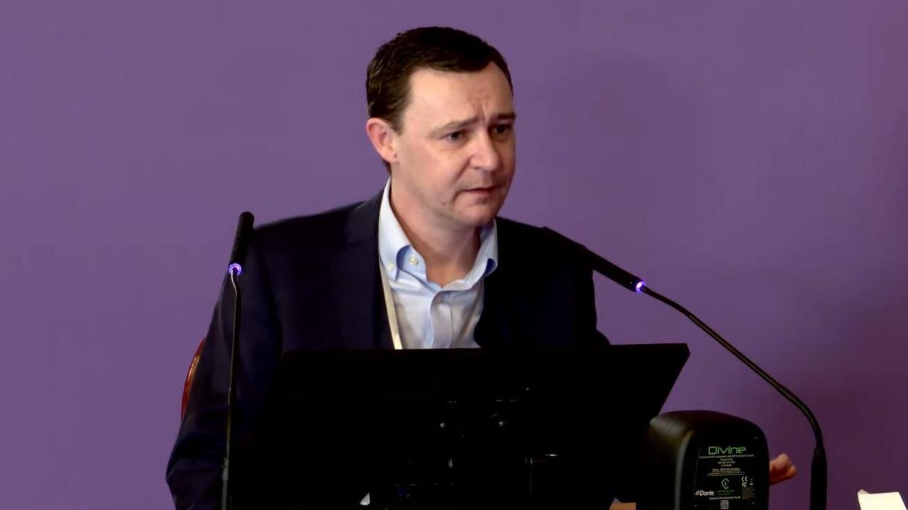 A man sits in front of an angles black screen and two microphones. He has short, dark hair, is clean shaven and is wearing a dark purple suit jacket with an open necked white shirt. The wall behind him is purple