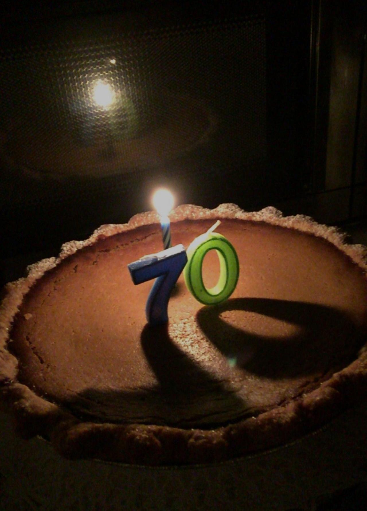 Birthday candles in the shape of the number 70 are alight on top of a pumpkin pie. They cast a shadow over the top of the pie