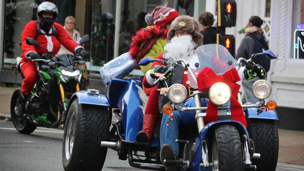 The bikers lining up the streets, dressed in Santa clothes. Their bikes are adorned with Christmas toys and tinsel. The biker in front has a red bow in front of his bike. The rider behind him is dressed as an elf.