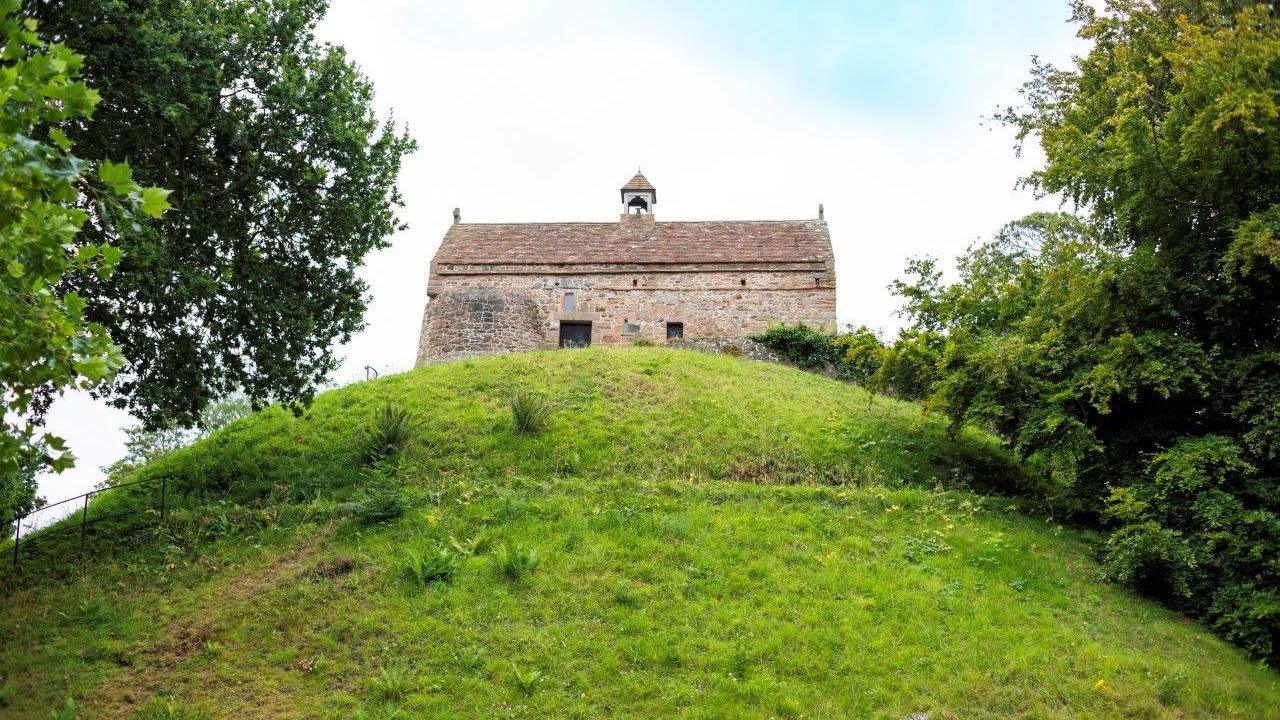 A brown brick building with a small bell tower in the middle of it on top of a green hill, two sets of trees surrounding either side, blue skies with white cloud