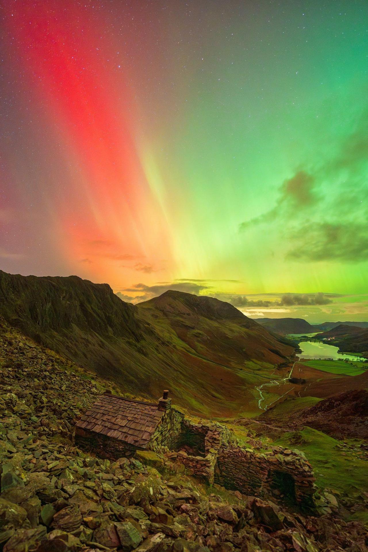 A stone shelter on a hillside. Above the building there is colourful curtain of light - to the left are red lights, while to the right are green lights.