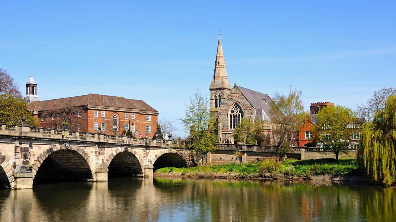 An arched stone bridge over a river with a church spire and a large brick building seen on the far side and the buildings and trees reflected in the water