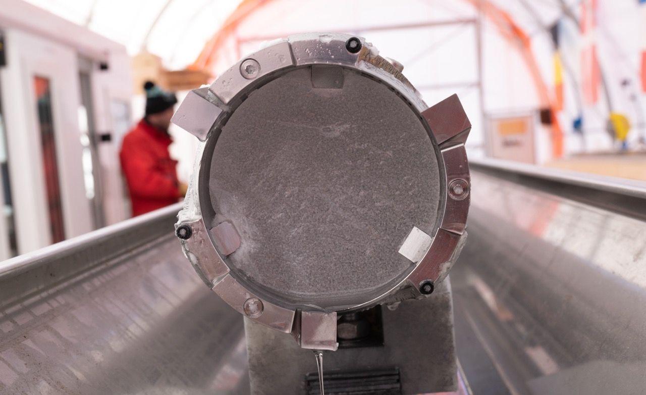 The end of an ice core suspended in a metal tube, inside a metal gulley. It is inside a large tent in Antarctica where the ice cores are being kept. On the left-hand side, there is a person wearing a bright red jacket and a bobble hat.