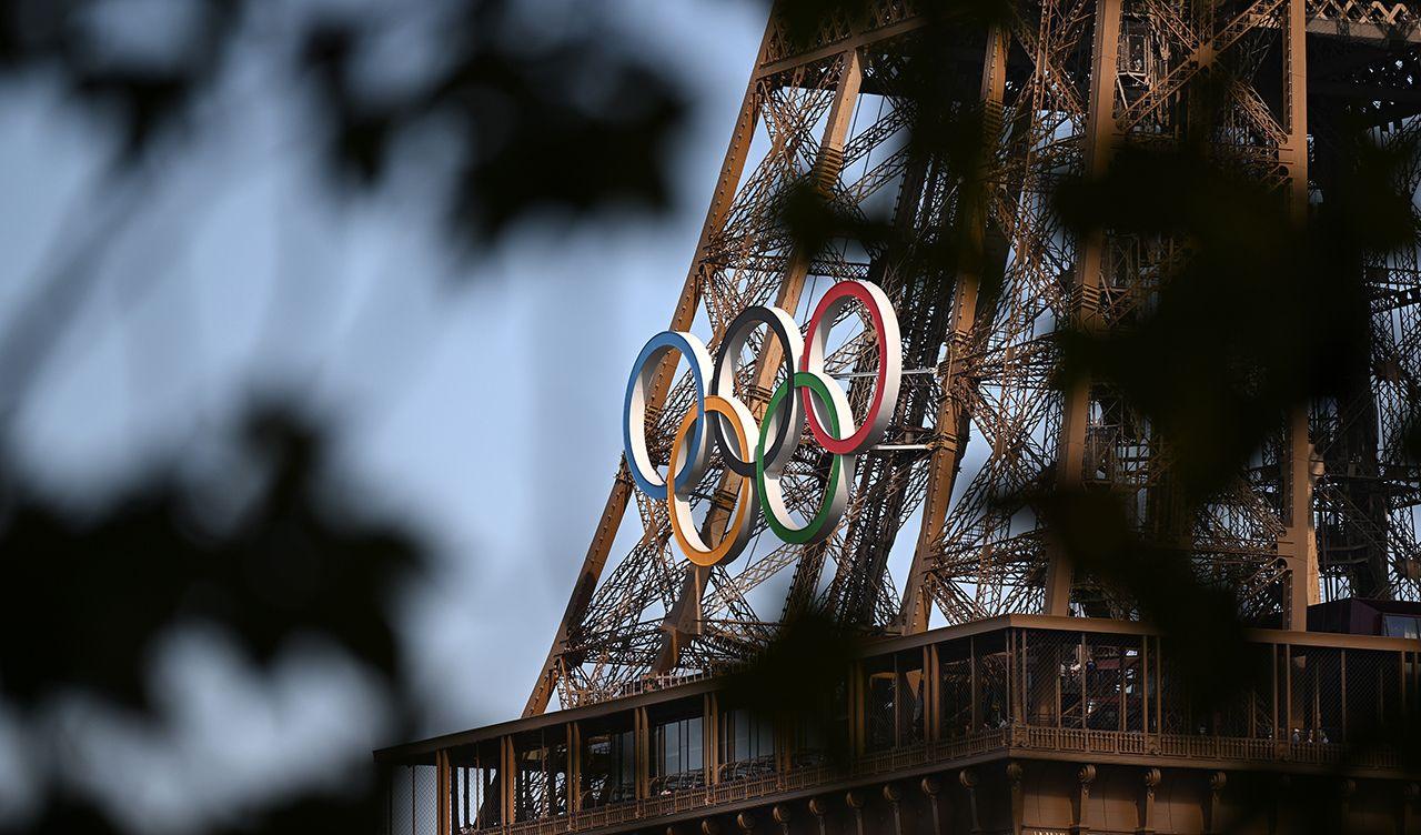 The olympic rings logo are seen attached to the front of the Eiffel tower through a tree with out-of-focus leaves in the foreground, in Paris on 26 August