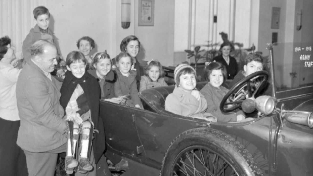 A black and white photo shows an old open top car with several children and teenagers sitting in it, smiling at the camera. A man stands next to the car and carried a child whose legs are both braced. The car is inside an office, with plants in the background.