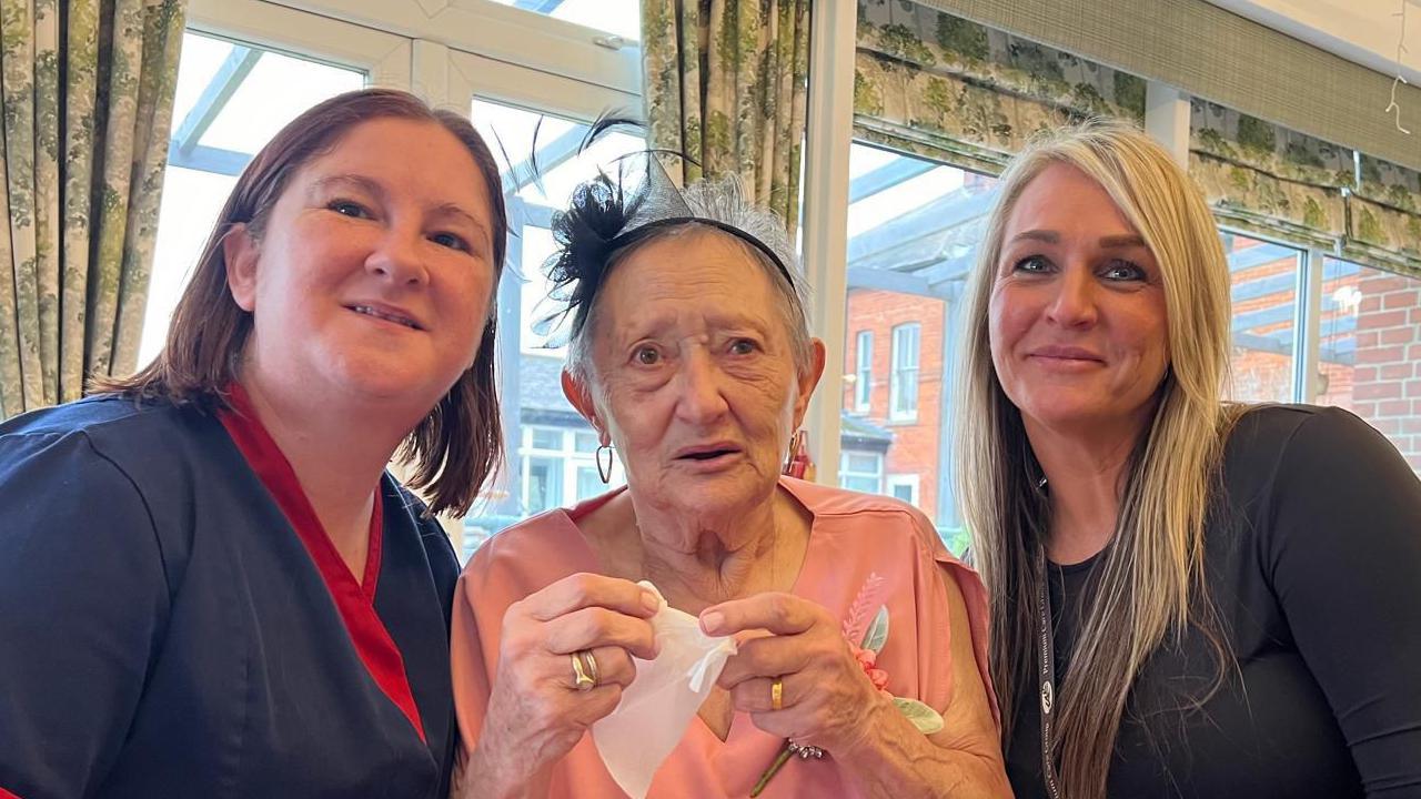 Care home staff member Charlie, Mrs Harris and care home manager Danielle Barham are pictured smiling at the camera while sitting next to each other. Charlie has shoulder length dark red hair and is wearing a care home uniform in navy. Ms Barham has long blonde hair and is wearing a black top.