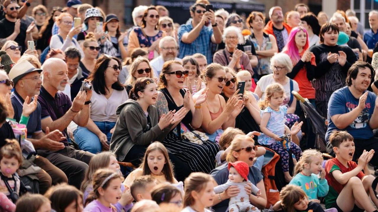 A large crowd of women, men and children applauds. Some are holding babies. They are mostly seated outside, on a bright and sunny day, with some people standing to the rear of the crowd. Some people are smiling, others are open-mouthed, some appear to be recording what they see on their phones and cameras.