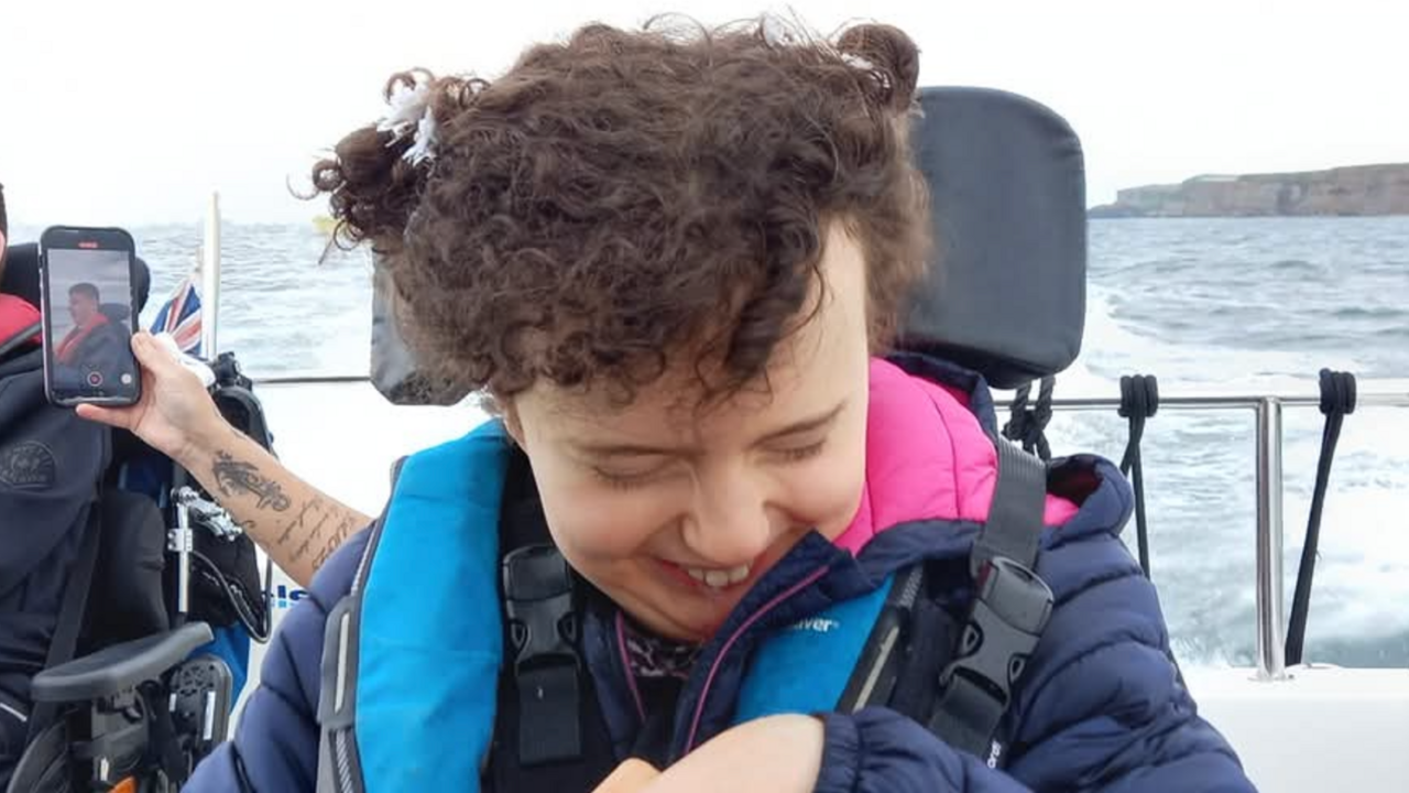 Amelie, who has curly dark hair tied up in small bunches, is wearing a navy blue puffer coat and is smiling. She's on a boat with water visible behind her.