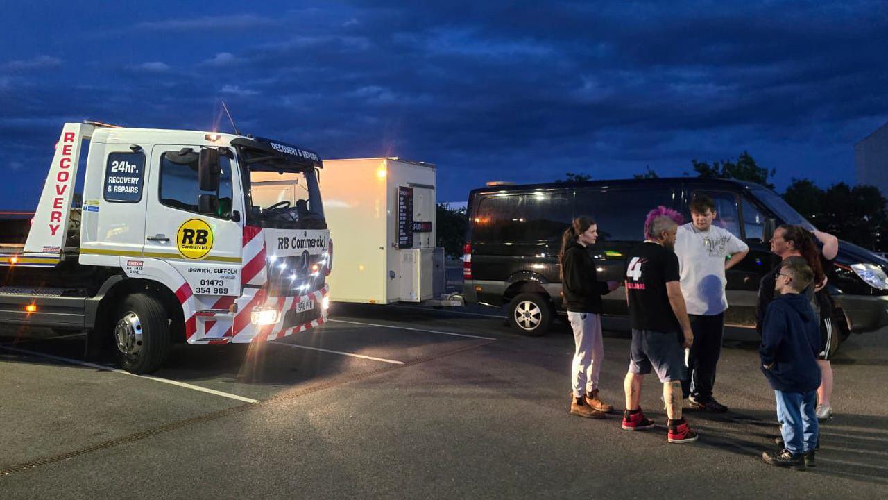 Spudman stands with a group of people in a car park talking to them. A white recovery vehicle can be seen parked next to a black van with a white street vendor attached to it.