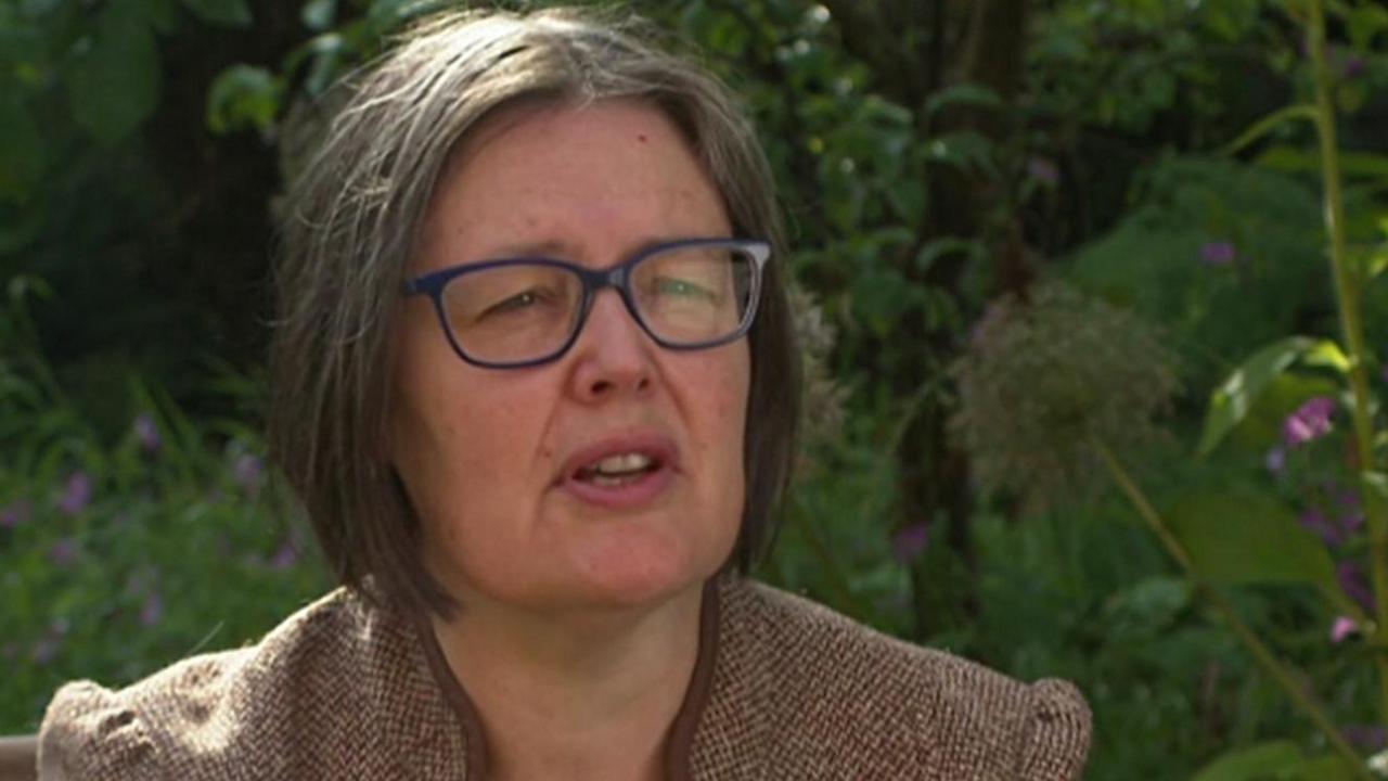 Alison Norris with chin-length brown hair in blue glasses wearing a brown blazer while sitting in a garden surrounded by greenery
