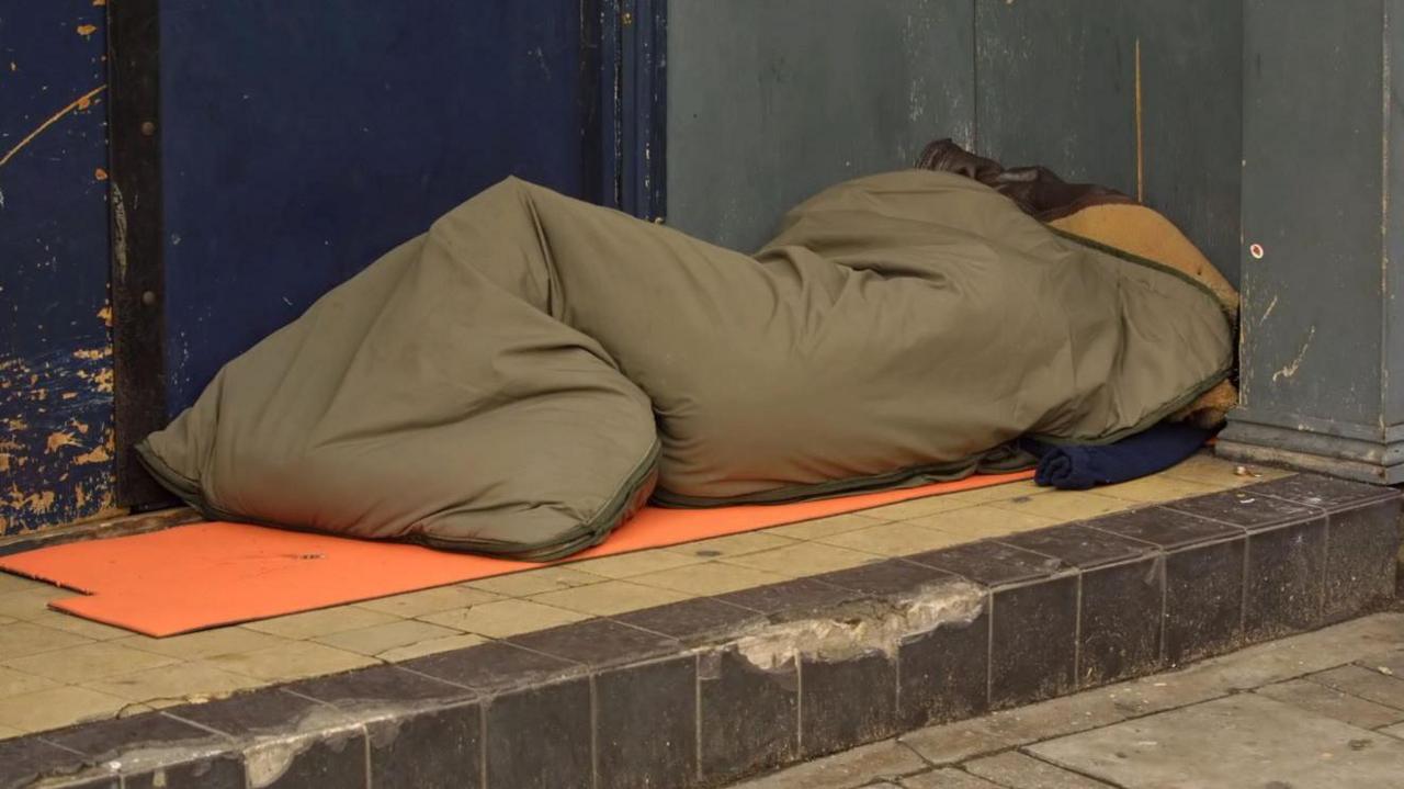 A khaki sleeping bag on a step outside a building with a blue door with someone appearing to be in it. An orange cardboard sheet is underneath the sleeping bag