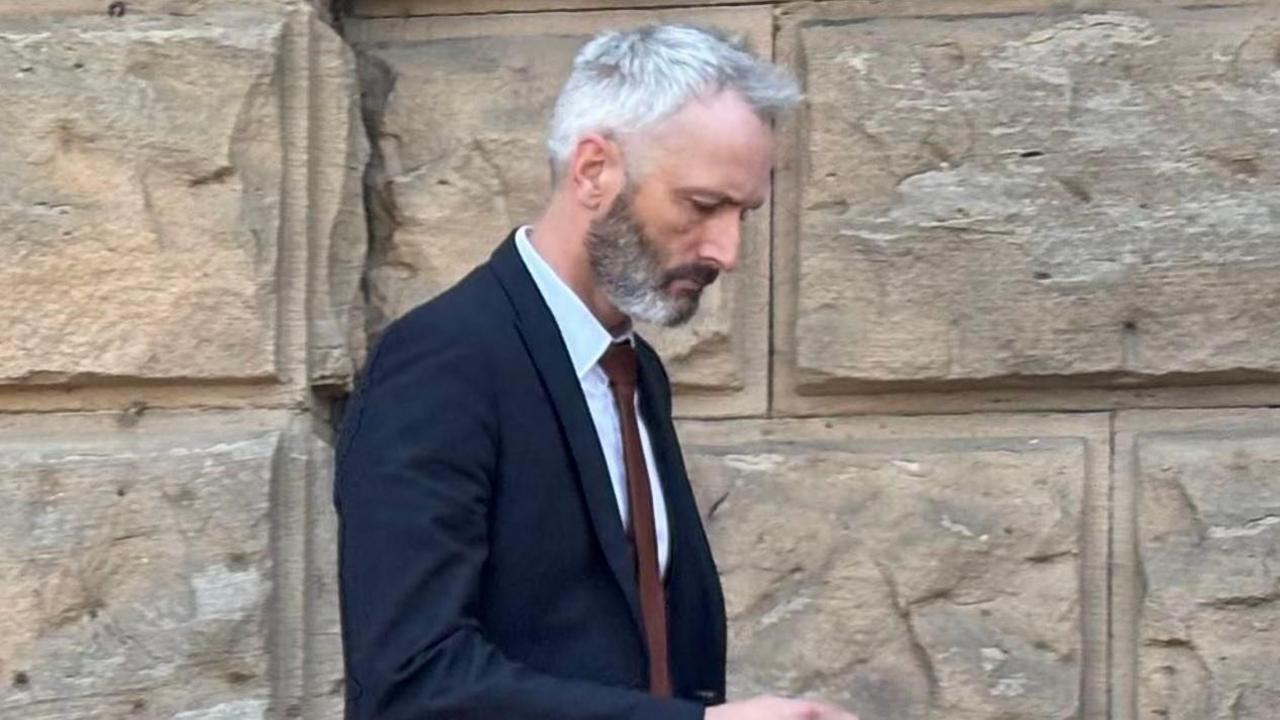 A side view of Lee Harshey-Jones leaving court. He wears a dark suit, white shirt and red tie and has his head down. 