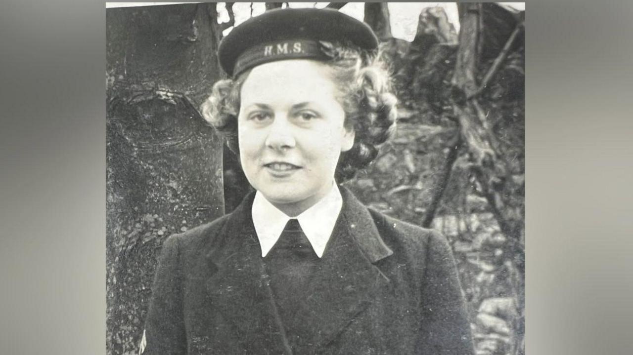 A black and white photo of a woman in uniform wearing a hat marked 'H.M.S.' stands outdoors with trees or foliage in the background. The photo was taken during the second world war