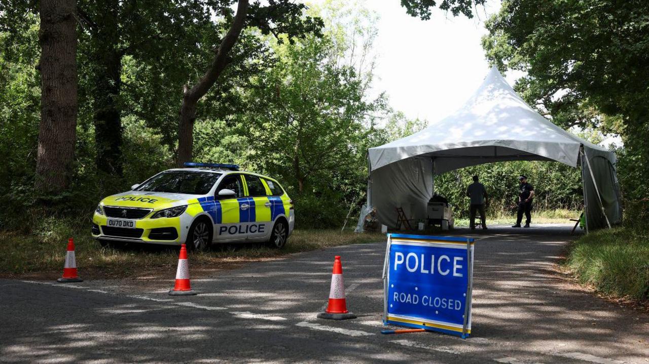 A police car is parked on the side of a road, blocked by traffic cones and a sign reading "Police road closed", in the hamlet of Dean in the Cotswolds. Behind there is a white tent, a police officer and a security patrol.