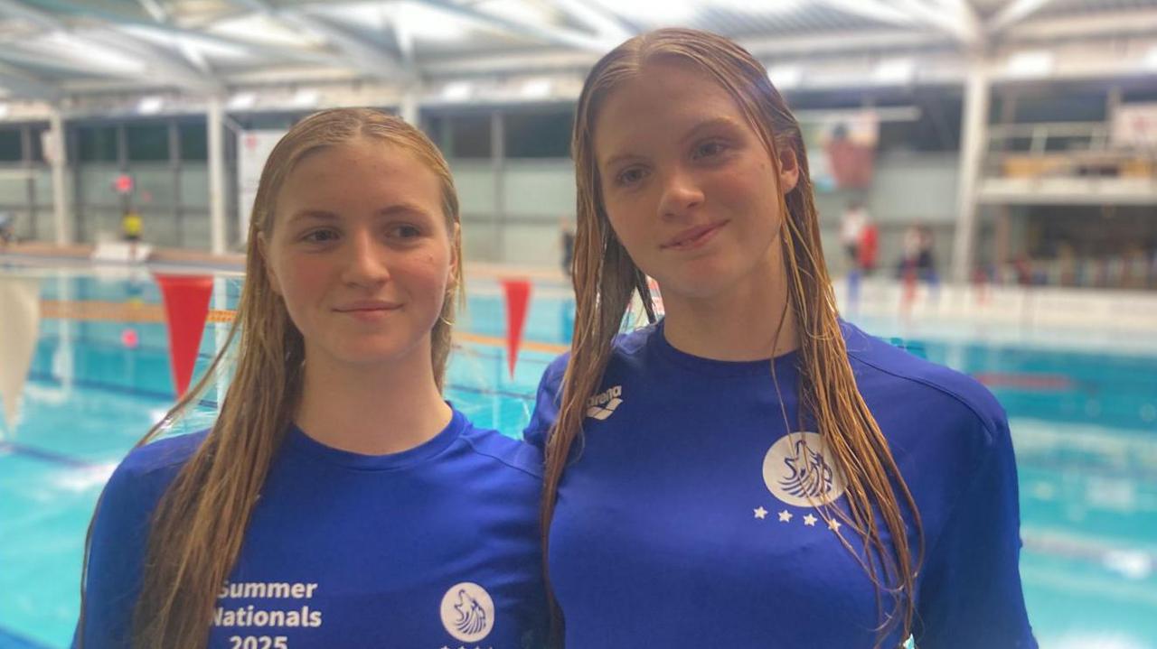 The sisters stand wearing blue tshirts in front a swimming pool. Their hair is damp and it looks like they've just finished a training session. 