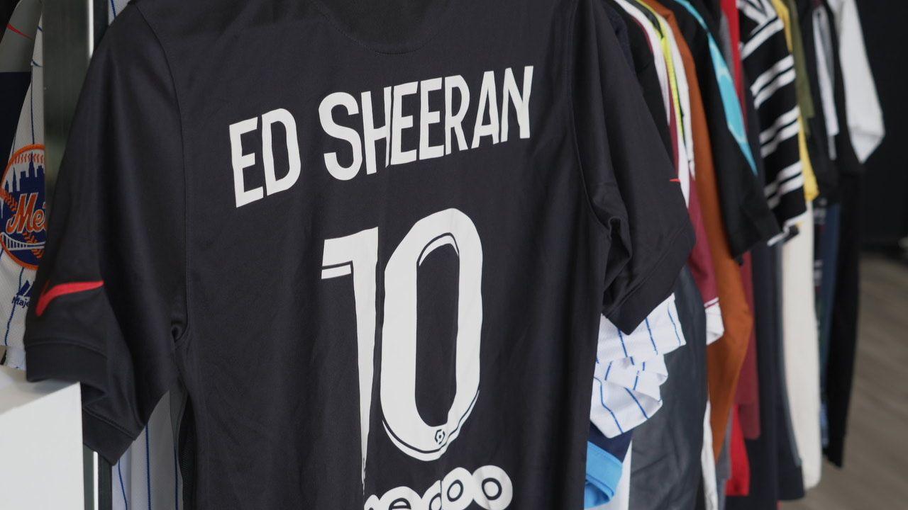 A football shirt with Ed Sheeran written on the back and the number 10. It is hanging on a rack along with several other tops owned by the singer. It is a black top and the writing is written in white.