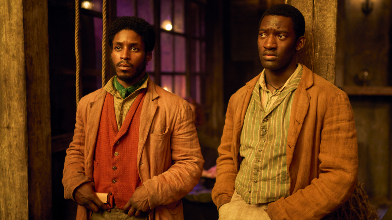 Francis Lovehall (left) Malachi Kirby (right) in costume as their characters Alec Munroe and Hezekiah Moscow. Both are wearing light brown jackets and colourful patterned waistcoats