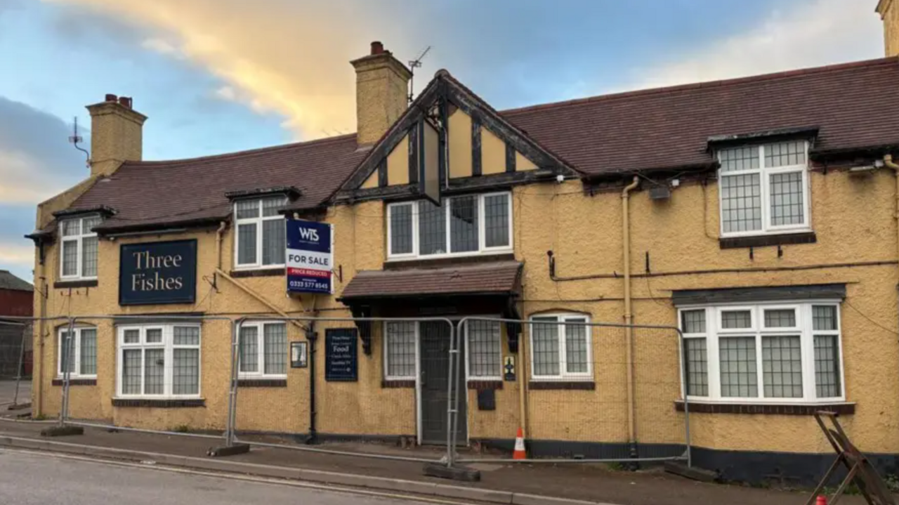 The front of a rendered light brown pub with the words Three Fishes on a dark blue sign and metal fencing around the front