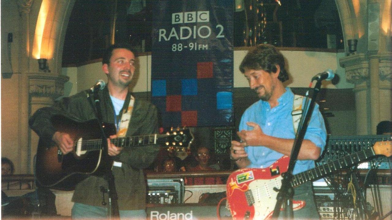 Musicians Paul Casey and Chris Rea share a BBC Radio 2 stage. A promo for radio 2 can be seen at the back of the stage, between Casey, who is on the left and wearing a green jacket, and Rea on the right, and in a blue shirt