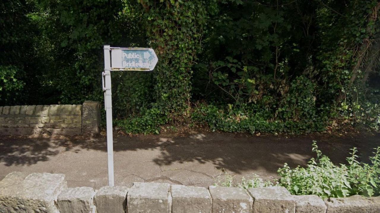 A blue sign with the words public bridleway in white in front of green foliage and above a stone wall