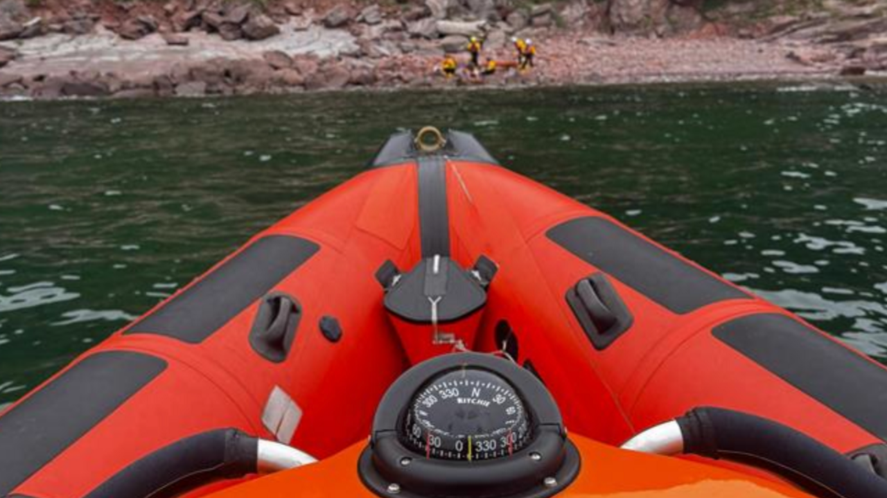The bow of an orange RIB at sea with a number of people on a beach.