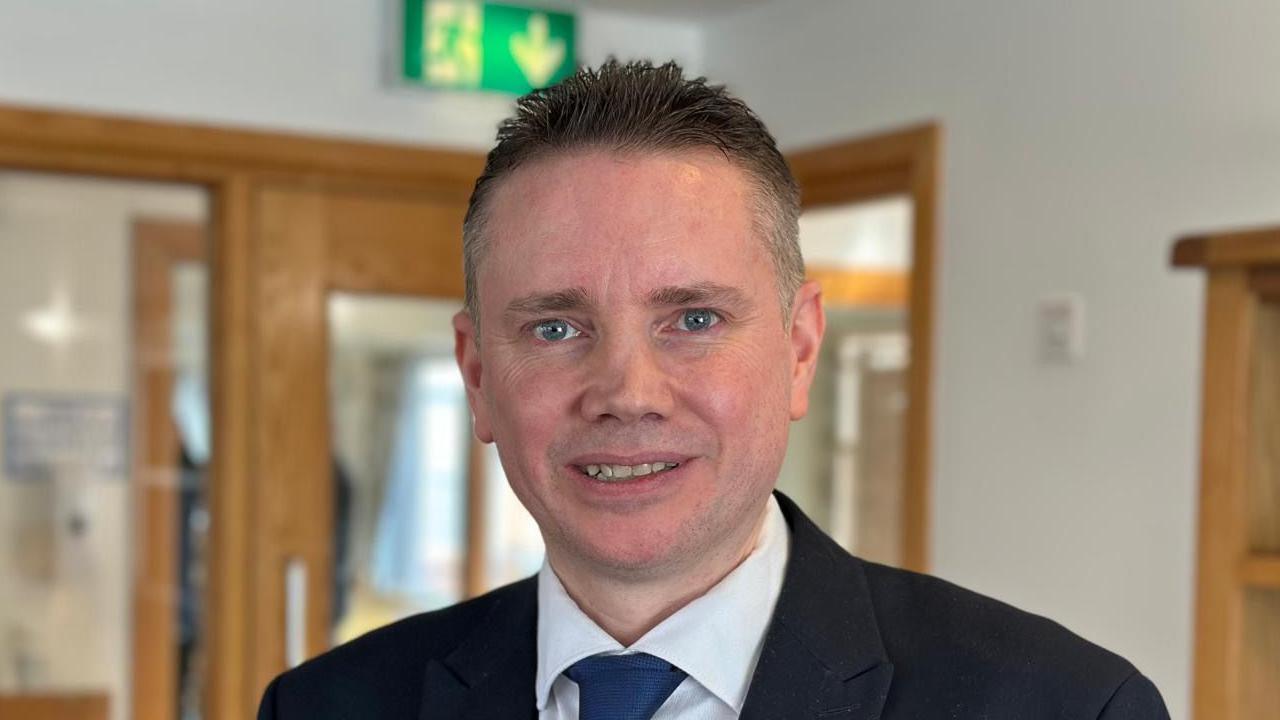 Chris Irwin has short dark hair, blue eyes and stubble. He is wearing a black blazer, white shirt and dark blue tie.