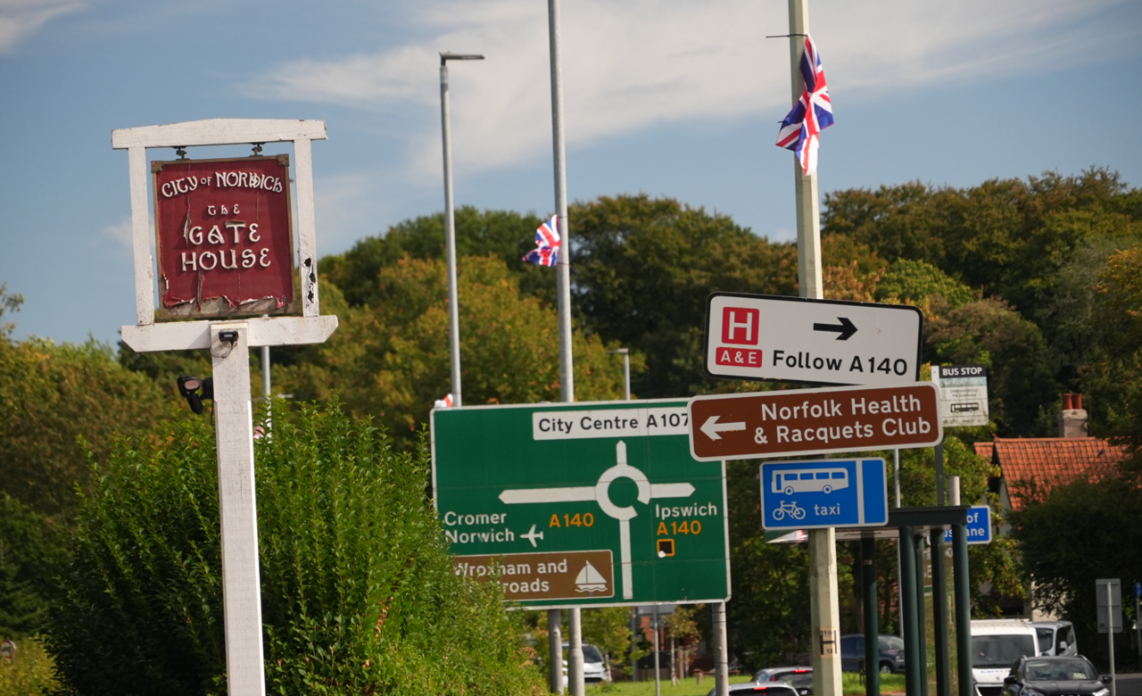 Two Union flags on lampposts at a roundabout. Road signs for a hospital, a bus line and a health club are on one of the lampposts. There is also a large green sign for the roundabout, indicating the city centre straight ahead, Cromer to the left and Ipswich to the right.