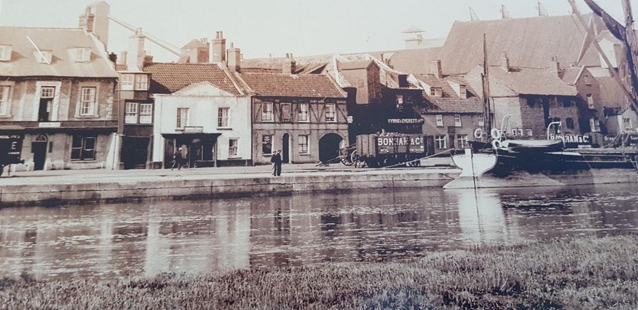 Image of Wells Harbour from around the time that French's fish and chip shop moved on to the waterfront. In the foreground is the water and then a line of shops and buildings along the quay. A boat is in the right hand side of the frame. 