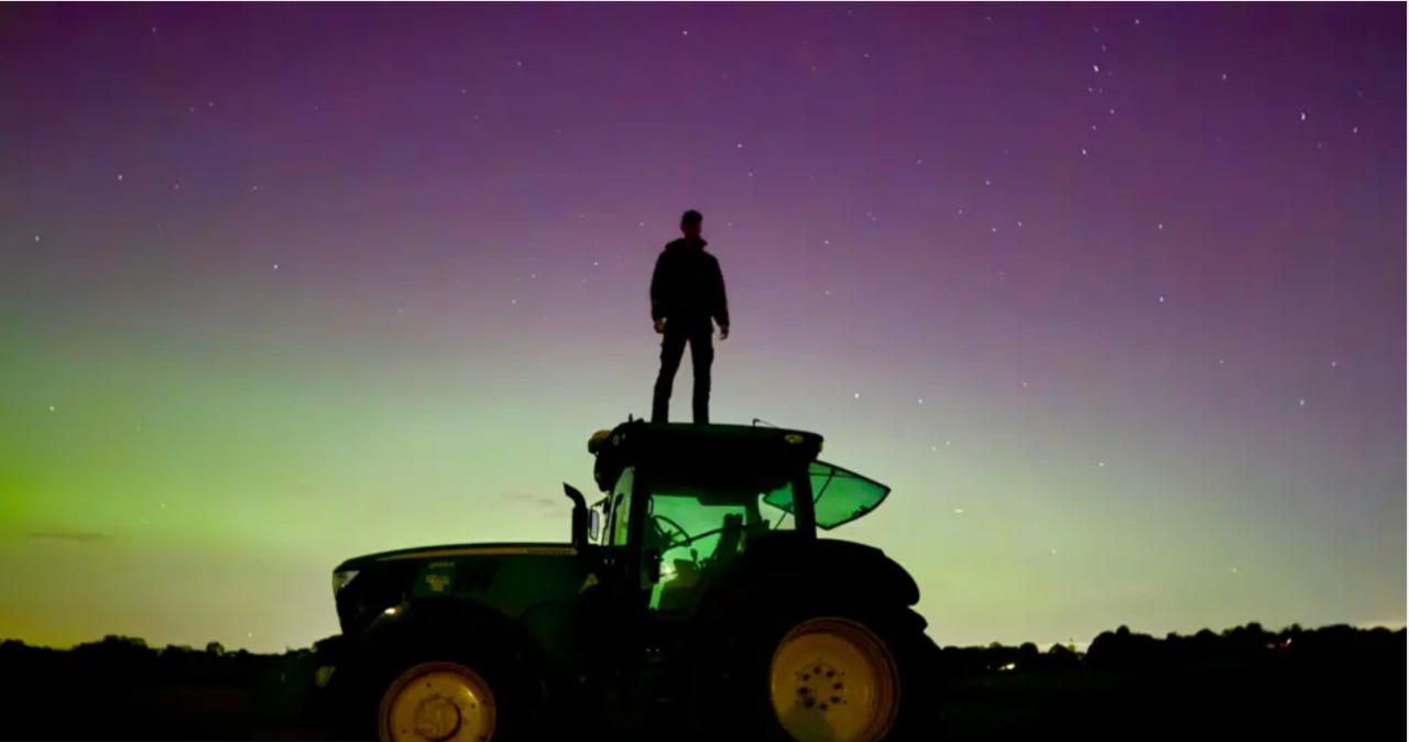 Northern lights fill the sky while a man can be seen standing on top of a tractor.