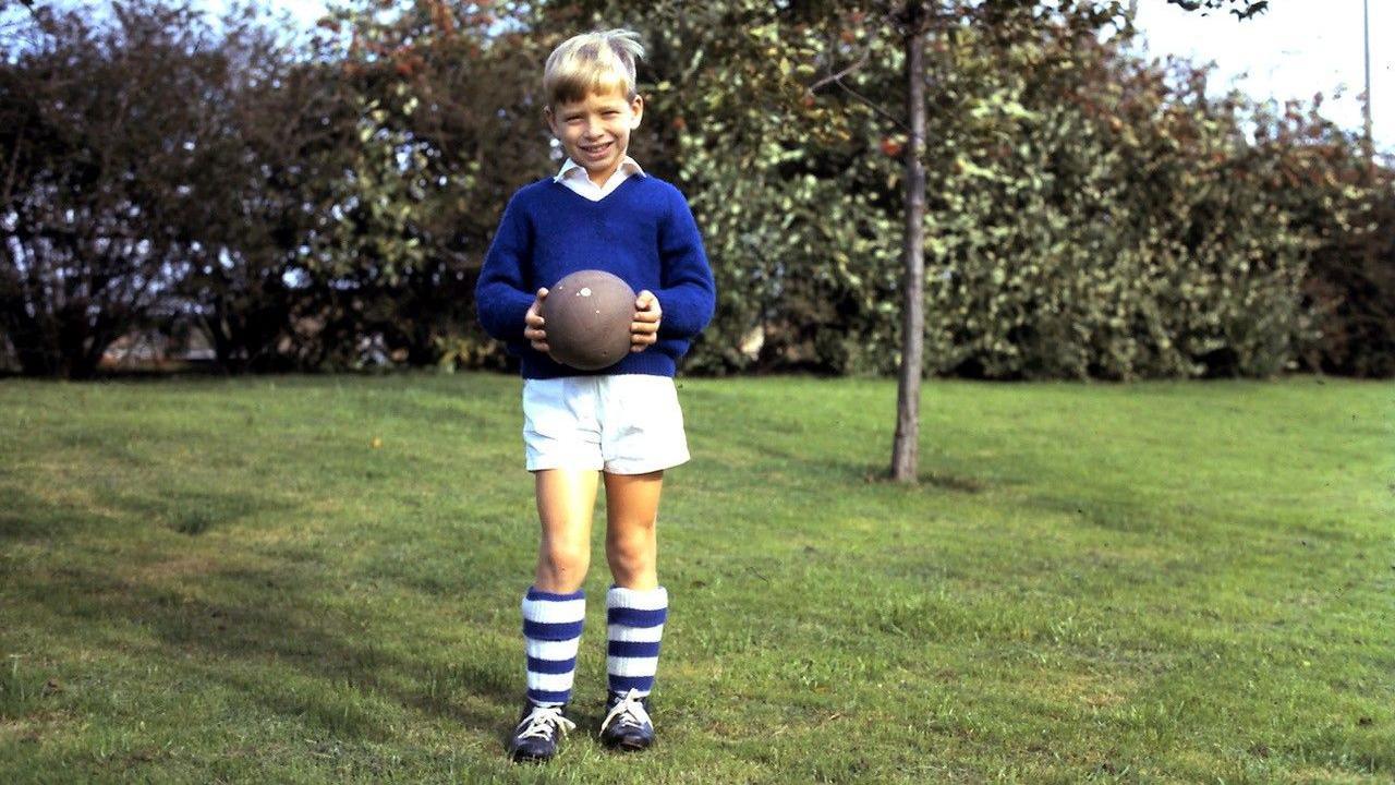 A boy wearing a dark blue jumper, white shorts and white and blue striped long socks. He is holding a ball and standing at a park.